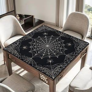 May include: A square black tablecloth featuring a white spiderweb pattern with multiple spider illustrations. The fabric covers the top surface of a wooden table, extending slightly over the edges, with the intricate web design centred.