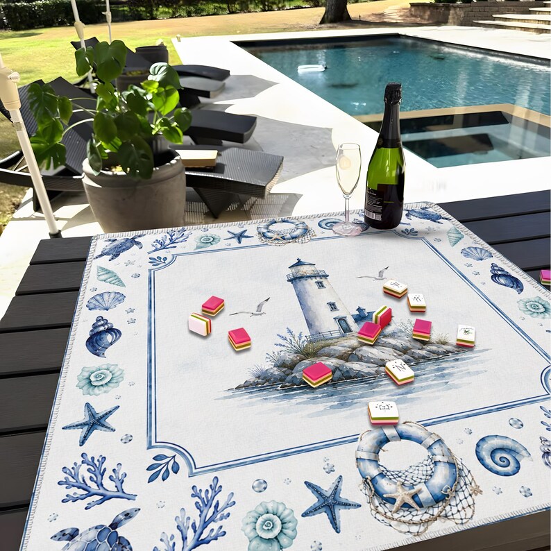 Puede incluir: Una mesa de tem&aacute;tica n&aacute;utica con un mantel individual blanco y azul con un faro, conchas marinas y estrellas de mar. Una botella de champ&aacute;n y una copa est&aacute;n sobre la mesa, junto con piezas de juego coloridas. La escena est&aacute; ambientada al aire libre, cerca de una piscina.