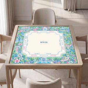 May include: A square mahjong mat featuring a cream-colored center with the blue monogram RWH. The border displays a vibrant tropical garden scene with pink flowers, green palm leaves, blue fountains, and lounge chairs, including game instructions.