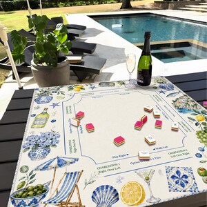 May include: A decorative game board with a coastal theme, featuring illustrations of olives, lemons, and a beach scene. The board has pink and white game pieces, a bottle of champagne, and a glass. The board is on a black table near a pool.