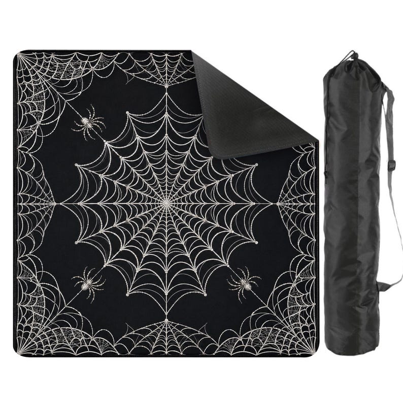 May include: A square black blanket featuring a white spiderweb pattern with small spiders, shown with one corner folded over to reveal a solid black underside. A matching black cylindrical carrying bag with a shoulder strap is positioned to the right.