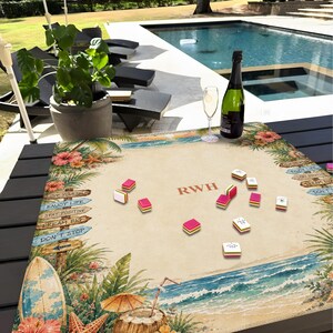 K&ouml;nnte beinhalten: Ein Mahjong-Set mit rosa und wei&szlig;en Spielsteinen auf einem Tisch mit Strandmotiv. Das Spielbrett zeigt eine tropische Strandszene mit Surfbrett, Meereswellen und dem Text "RWH". Eine Flasche Champagner und ein Glas stehen ebenfalls auf dem Tisch.