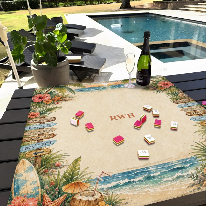 K&ouml;nnte beinhalten: Ein Mahjong-Set mit rosa und wei&szlig;en Spielsteinen auf einem Tisch mit Strandmotiv. Das Spielbrett zeigt eine tropische Strandszene mit Surfbrett, Meereswellen und dem Text "RWH". Eine Flasche Champagner und ein Glas stehen ebenfalls auf dem Tisch.