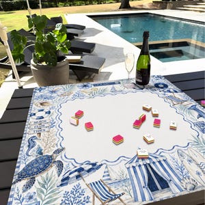 May include: A beach-themed game board with a white center and a blue and white border featuring illustrations of a turtle, seahorse, and beach chairs. The board has the text "Beach Club Tides" and is surrounded by game pieces, a bottle of champagne, and a glass.