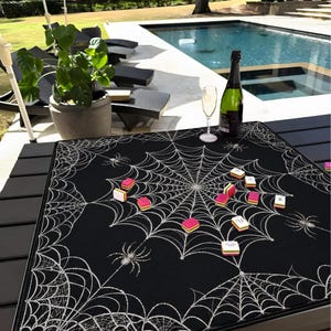 May include: A square black game mat featuring a white spiderweb pattern with several spiders and scattered colourful mahjong tiles. The mat is placed on a dark slatted table, with a bottle of champagne and a glass visible in the background.