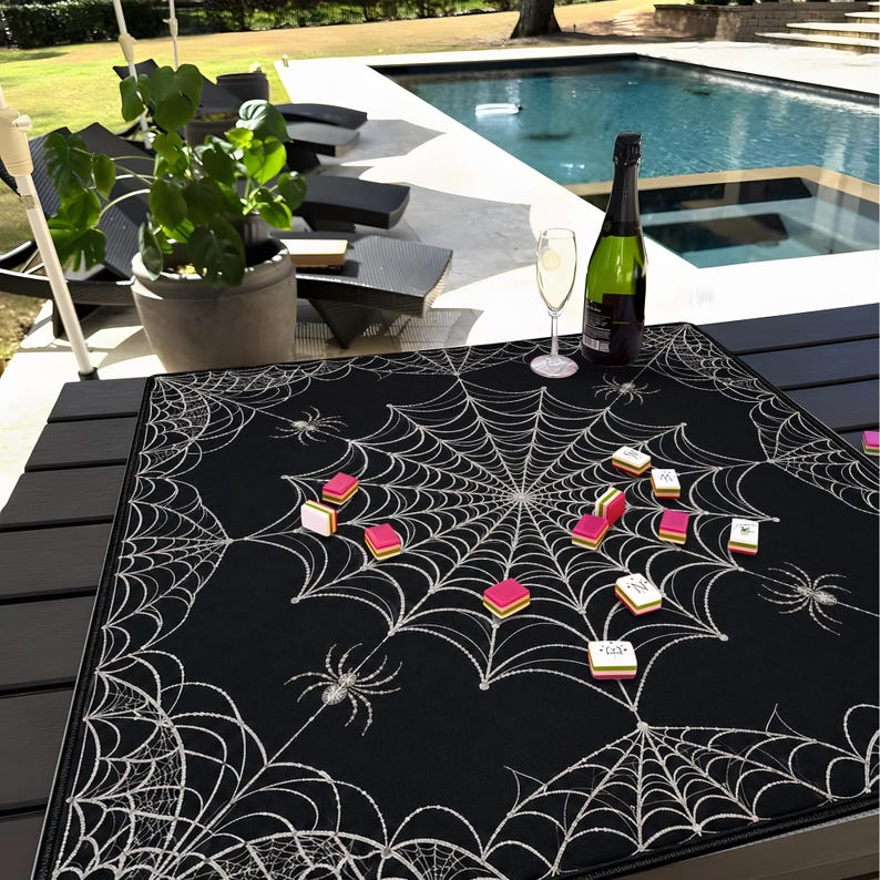May include: A square black game mat featuring a white spiderweb pattern with several spiders and scattered colourful mahjong tiles. The mat is placed on a dark slatted table, with a bottle of champagne and a glass visible in the background.