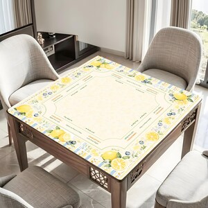 Pu&ograve; includere: Un tavolo in legno con un piano decorativo a motivi di limoni e mirtilli. Il tavolo ha uno sfondo giallo chiaro con un bordo di limoni, mirtilli e testo. Il tavolo &egrave; circondato da tre sedie grigie.