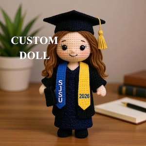 Puede incluir: Muñeca de ganchillo hecha a mano con birrete y toga de graduación. La muñeca tiene cabello castaño y lleva una banda azul y amarilla con el texto "SJSU" y "2026". El texto "CUSTOM DOLL" se muestra en la imagen.