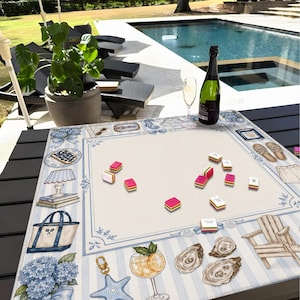 May include: A decorative game board with a blue and white striped border, featuring illustrations of beach-themed items. Pink and yellow game pieces are scattered across the center. A bottle of champagne and a glass are on the table.