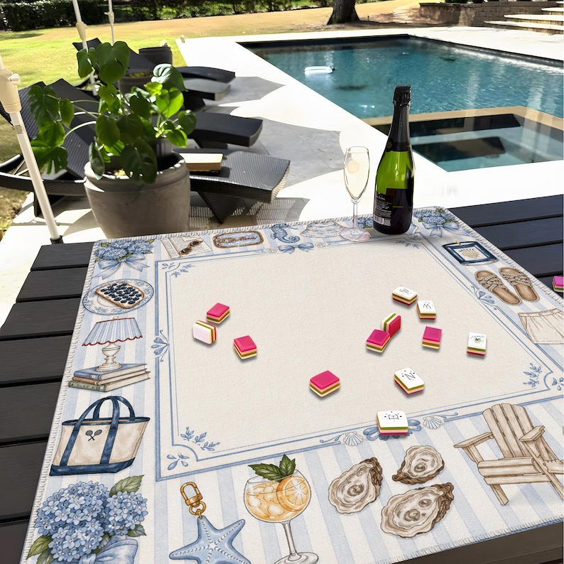 May include: A decorative game board with a blue and white striped border, featuring illustrations of beach-themed items. Pink and yellow game pieces are scattered across the center. A bottle of champagne and a glass are on the table.