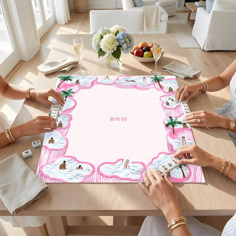 May include: A rectangular mahjong mat featuring a light pink center with the letters RWH in the middle. The border displays a repeating pattern of pink-rimmed swimming pools with blue water, palm trees, and small figures, with mahjong tiles.