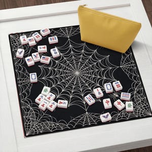 May include: A black square mat featuring a white spiderweb pattern, scattered with white Mahjong tiles displaying various symbols, numbers, and American flag designs, accompanied by a solid yellow zippered storage pouch.