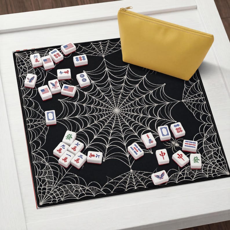May include: A black square mat featuring a white spiderweb pattern, scattered with white Mahjong tiles displaying various symbols, numbers, and American flag designs, accompanied by a solid yellow zippered storage pouch.