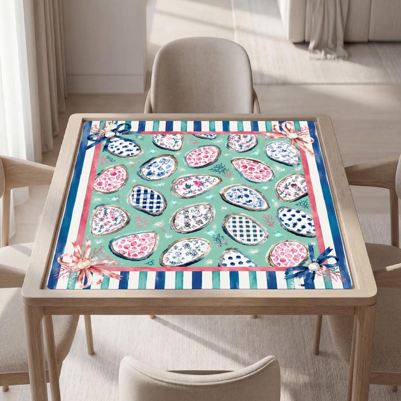 May include: A square tablecloth featuring a light green background with a repeating pattern of oyster shells decorated in various blue and pink floral, plaid, and polka dot prints, framed by a border of blue stripes and pink ribbons at corners.