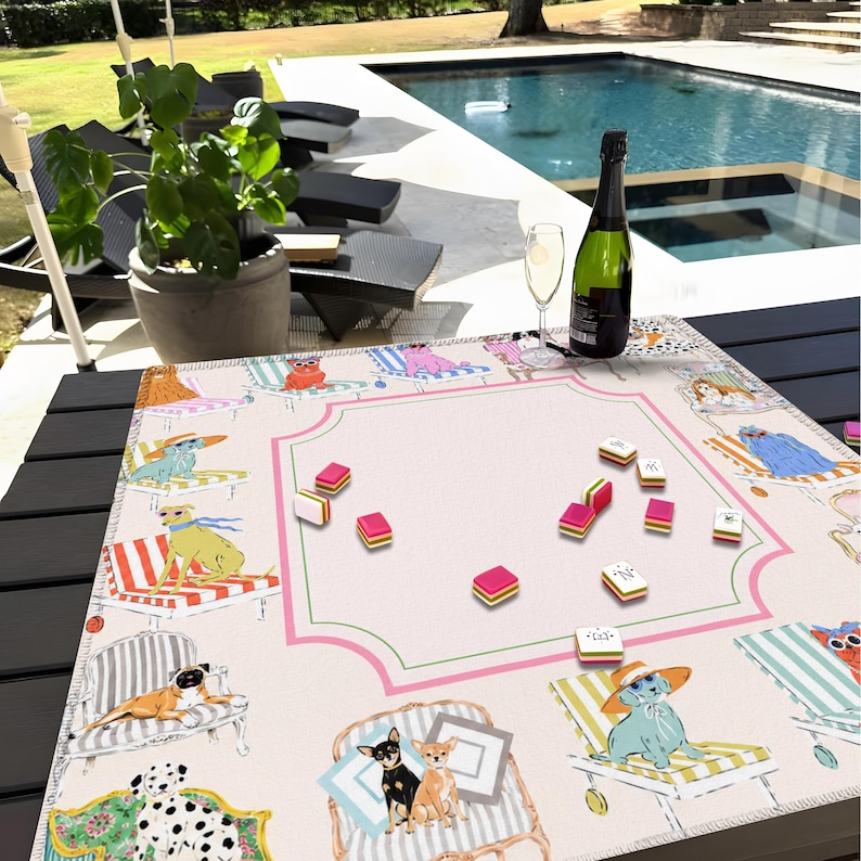 May include: A light pink Mahjong game mat featuring illustrations of dogs and cats relaxing on beach chairs. The mat has a pink and green border, with Mahjong tiles scattered across the surface. A bottle of champagne and a glass are on the table.