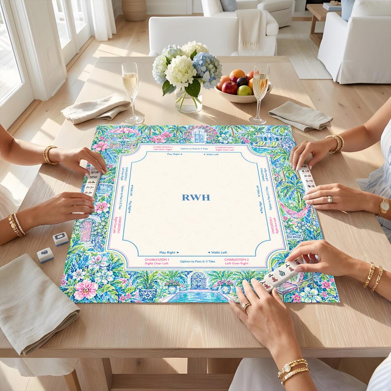 May include: A square mahjong mat featuring a vibrant floral and tropical garden border in shades of blue, pink, and green. The center is cream-colored with blue lines, game instructions, and the monogram RWH printed in the middle.