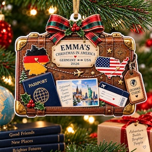 May include: A brown suitcase-shaped Christmas ornament featuring a red, black, and gold map of Germany and a red, white, and blue map of the USA. It includes a blue passport, a postcard with the Statue of Liberty, and text reading EMMA'S CHRISTMAS IN AMERICA GERMANY USA 2026.