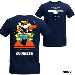 Peut inclure: T-shirts bleu marine avec un motif Guinness. L'un pr&eacute;sente une illustration de toucan avec une pinte de Guinness, des citrons et le texte "It's a lovely day for a Guinness". L'autre a un graphique de pinte de Guinness et du texte.