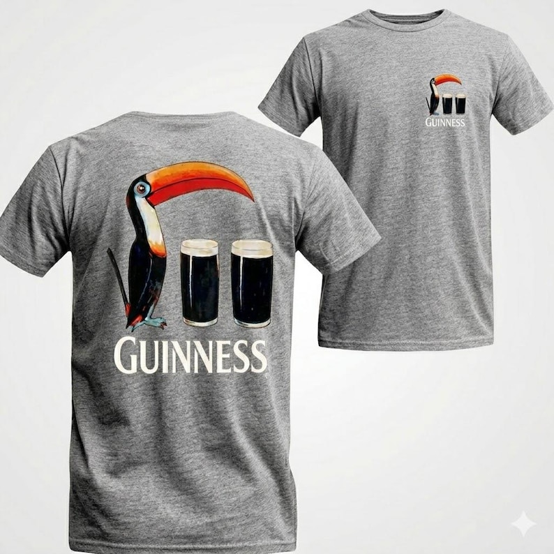 May include: Gray t-shirts with a Guinness theme. One shirt features a toucan with a large orange beak and two pints of dark beer, with the Guinness logo below. The other shirt has a smaller version of the design on the pocket.