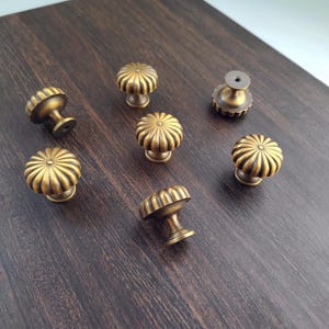 May include: A selection of antique brass cabinet knobs. Each knob has a ribbed, pumpkin-shaped design with a short stem and a round base. The knobs are arranged on a dark wood surface, highlighting their warm, metallic colour.