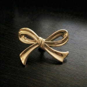Pode incluir: Um broche dourado em forma de laço. O broche tem um design detalhado com um nó amarrado no centro e laços de fita fluindo. O laço está sobre um fundo escuro.