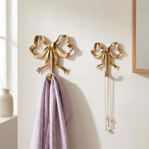 Peut inclure: Deux patères murales dorées en forme de nœud papillon. La plus grande accroche une écharpe lavande à motifs cachemire. La plus petite accroche un collier doré avec un pendentif clé et perle. Sur un mur blanc.