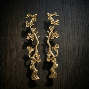 Puede incluir: Dos tiradores dorados para armarios con forma de ramas florecidas. Los tiradores presentan pequeñas flores y corteza texturizada, sobre un fondo de madera oscura. El diseño evoca una estética natural y orgánica.