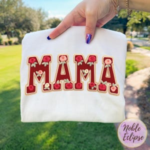 May include: White sweatshirt with the word "MAMA" embroidered in red with a gold outline. The letters are decorated with red and pink roses. The sweatshirt is held up by a hand with blue nail polish.