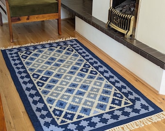 May include: A rectangular blue rug with a geometric diamond pattern in shades of blue and white. The rug has a dark blue border and fringe along the short edges. The rug is placed on a wooden floor.