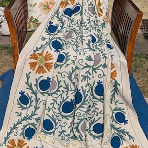 Könnte beinhalten: Cremefarbene Decke mit detailliertem Stickdesign. Die Decke zeigt blaue, orangefarbene und graue Blumen- und Granatapfelmotive mit grünen Ranken und einem Fransenrand. Die Decke ist über einen Holzstuhl drapiert.
