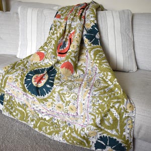 Puede incluir: Una manta decorativa con un estampado floral en verde oliva, azul marino, melocotón y crema. Está colocada sobre un sofá gris con cojines a rayas y lisos. El diseño de la manta presenta grandes motivos florales estilizados.