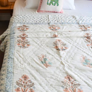 Könnte beinhalten: Eine weiße, gesteppte Tagesdecke mit einem sich wiederholenden floralen Blockdruckmuster in Pfirsich- und gedämpften Blautönen. Die Decke hat einen breiten Rand mit einem kleineren Blumenmotiv und eine rautenförmige Steppung auf der Oberfläche.