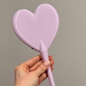 May include: A light purple hand mirror in the shape of a heart. The mirror has a long, cylindrical handle. The background is a solid, neutral gray.