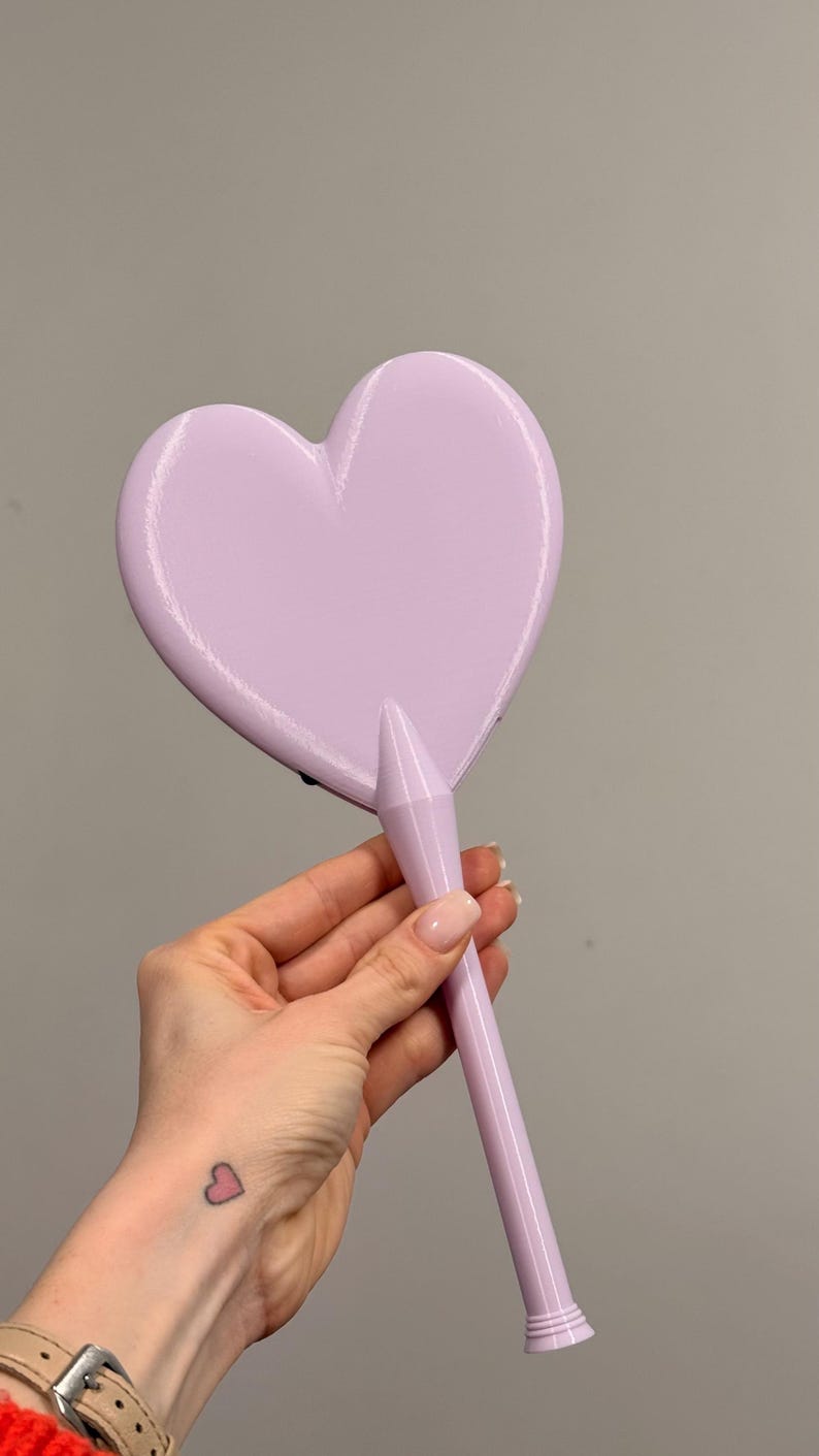 May include: A light purple hand mirror in the shape of a heart. The mirror has a long, cylindrical handle. The background is a solid, neutral gray.