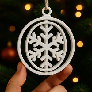Könnte beinhalten: Weißer Schneeflocken-Weihnachtsschmuck. Der Schmuck zeigt ein detailliertes Schneeflocken-Design in einem kreisförmigen Rahmen, der an einer natürlichen Schnurschlaufe aufgehängt ist. Der Hintergrund ist mit Weihnachtsbaumlichtern verschwommen.