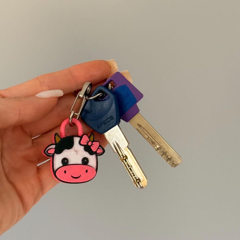 May include: A set of keys with a pink cow-shaped keychain. The keychain has a black and white cow face with a pink bow. The keys have blue and purple plastic heads.