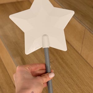 May include: A white star-shaped wand with a gray handle. The wand is held in a hand, with a small heart tattoo visible on the wrist. The wand is a whimsical accessory.