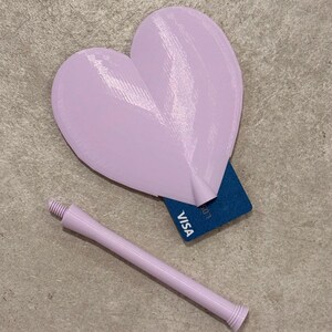 May include: A light purple heart-shaped object with a matching handle, possibly a bottle stopper. A blue Visa card is partially visible behind the heart. The objects are on a light gray surface.
