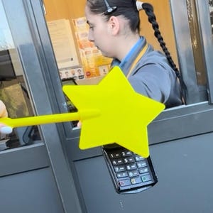 May include: A yellow star-shaped wand is held over a black payment terminal. The terminal has a keypad with numbers and symbols. The wand is being used at a drive-through window.