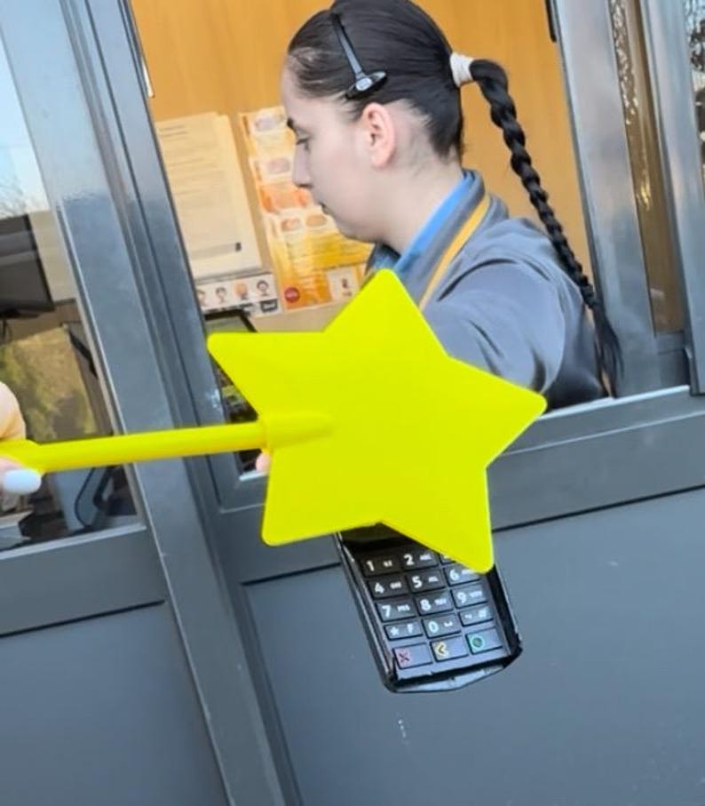 May include: A yellow star-shaped wand is held over a black payment terminal. The terminal has a keypad with numbers and symbols. The wand is being used at a drive-through window.