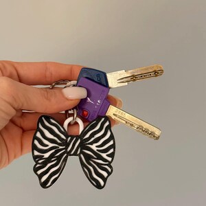 May include: A keychain featuring a black and white zebra-striped bow charm attached to a metal ring. Two keys are also attached to the ring: one with a blue plastic head and one with a purple plastic head labeled Mul-T-Lock with the number 2885.