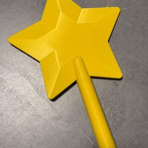May include: A bright yellow star-shaped object with a smooth, faceted surface and a cylindrical handle attached to the bottom point. The item features a distinct geometric design with sharp angles and a uniform, solid colour finish throughout.