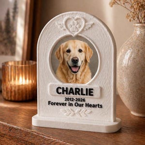 May include: A white memorial plaque with a photo of a golden retriever named Charlie. The plaque has a heart and paw print design at the top, with the name and dates below the photo. The text reads "Forever in Our Hearts."
