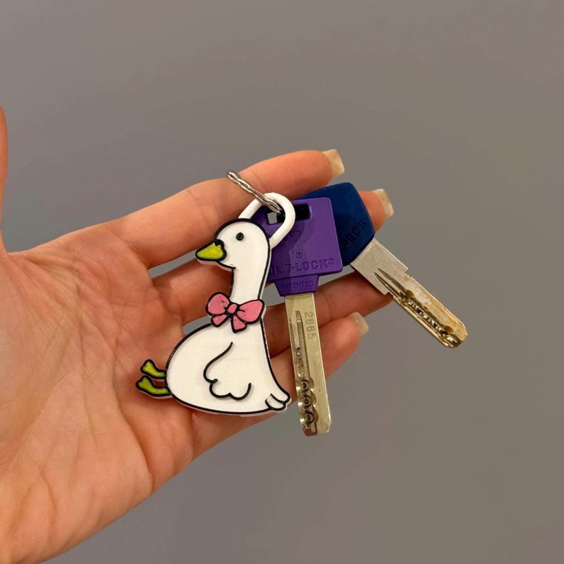 May include: A white enamel keychain shaped like a goose with a pink bow, a yellow beak, and green feet, attached to a silver metal ring holding two keys, one with a purple plastic head and one with a silver metal head.