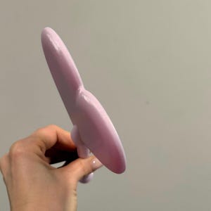 May include: A light purple, elongated object with a rounded tip and a bulbous base. The object is held in a hand against a plain background. The object appears to be made of a smooth material.