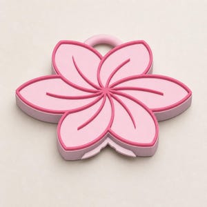 May include: A pink flower-shaped coaster with a loop for hanging. The coaster has a light pink base with a darker pink outline and petal details. The design is a stylized floral shape.