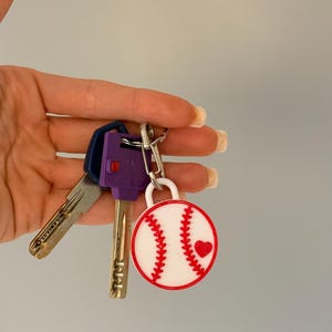 May include: A circular baseball keychain featuring a white background with red stitching and a small red heart accent, attached to a silver metal ring holding two keys, one with a purple plastic head and one with a standard metallic finish.