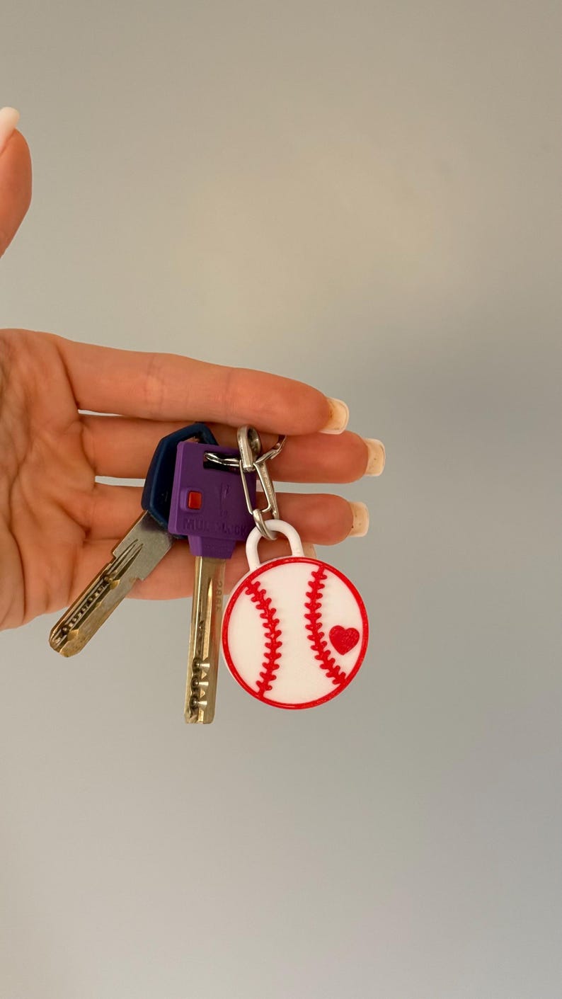 May include: A circular baseball keychain featuring a white background with red stitching and a small red heart accent, attached to a silver metal ring holding two keys, one with a purple plastic head and one with a standard metallic finish.