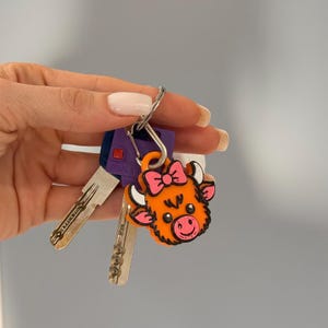 May include: A set of keys with a decorative orange cow-shaped keychain. The keychain features a pink bow and a smiling face. The keys are silver and purple.