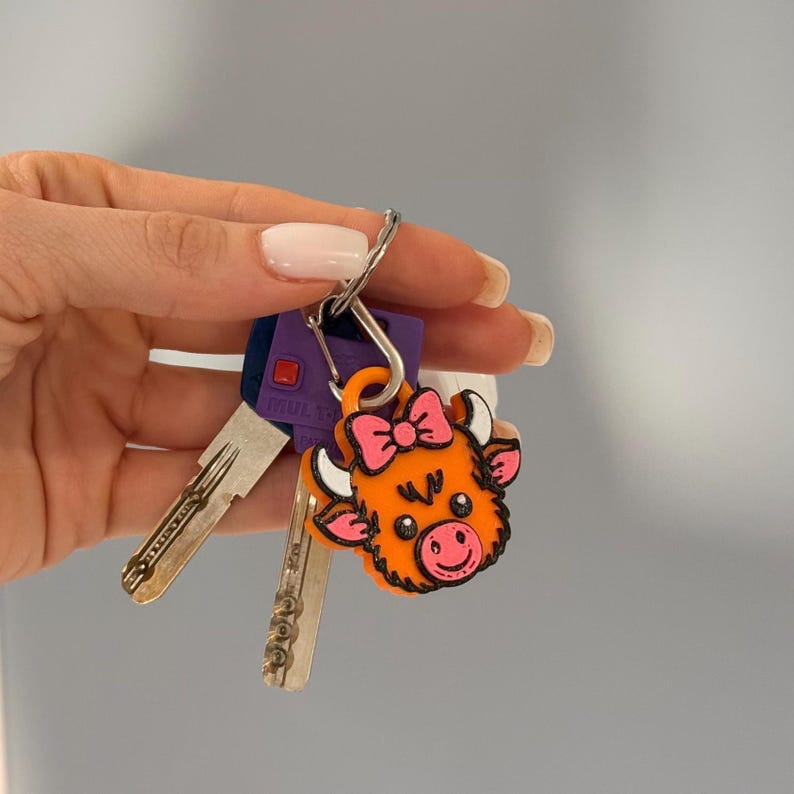 May include: A set of keys with a decorative orange cow-shaped keychain. The keychain features a pink bow and a smiling face. The keys are silver and purple.